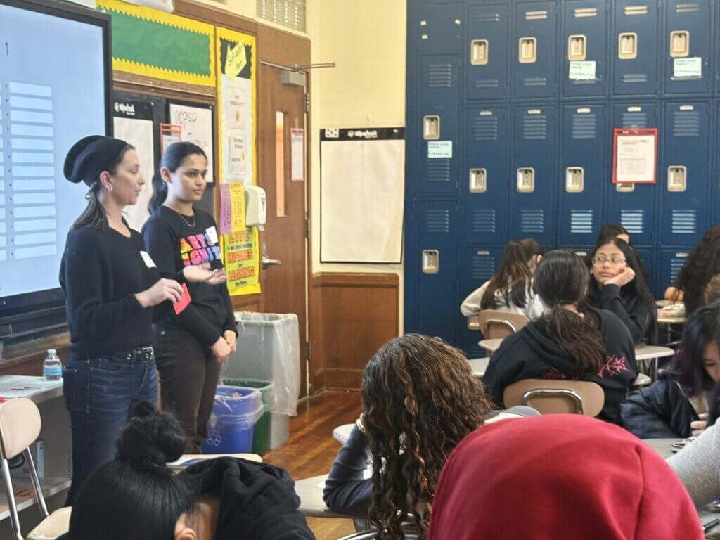 Two women stand at the front of a classroom speaking to students seated at desks. Lockers line the back wall, and a large screen with charts is visible on the left.