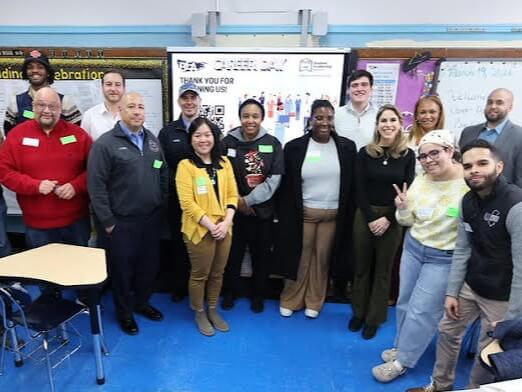 A group of people stands together in a classroom, posing for a photo in front of a board and banner, some smiling and wearing name tags.