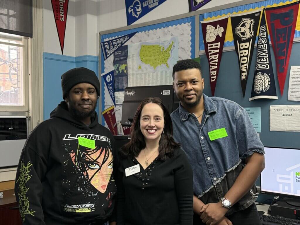 Three adults stand together in a classroom decorated with college pennants, a map, and educational posters.