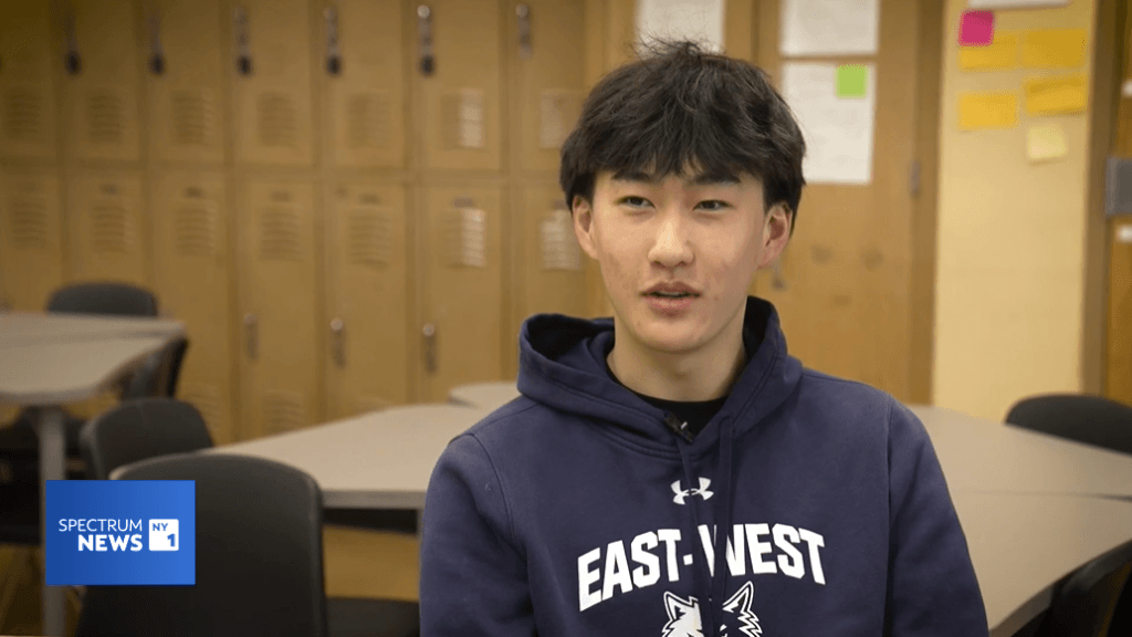 A student wearing an EAST-WEST hoodie sits in a classroom with beige lockers and tables in the background. The Spectrum News 1 logo appears in the corner for the sports scholarship news feature