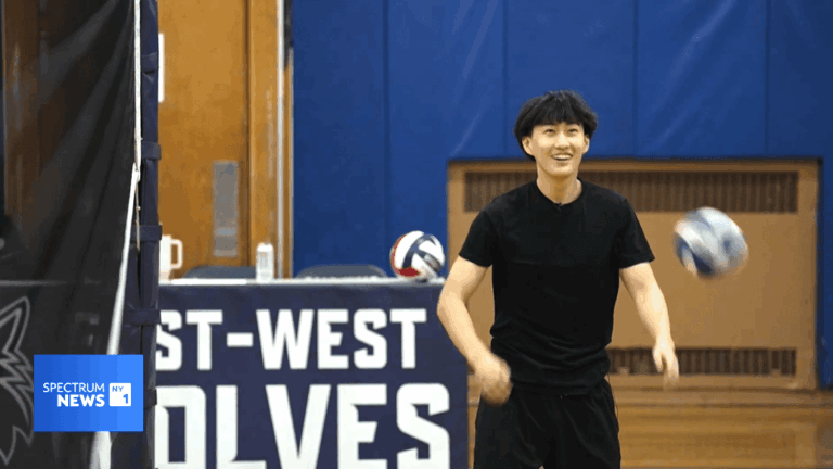 A student in black athletic clothing stands in a gym near a volleyball net, posing for a sports scholarship news feature