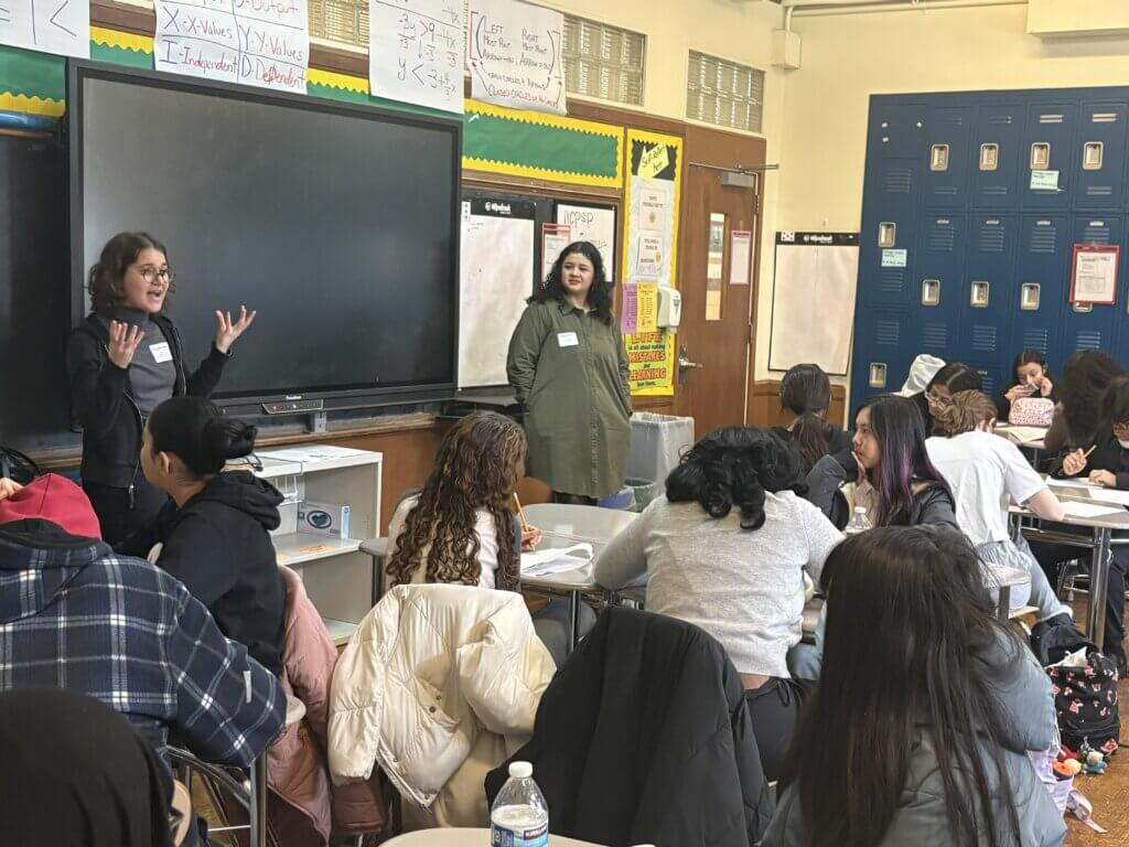 Two women stand at the front of a classroom speaking to students seated at desks, with educational posters and lockers visible in the background.