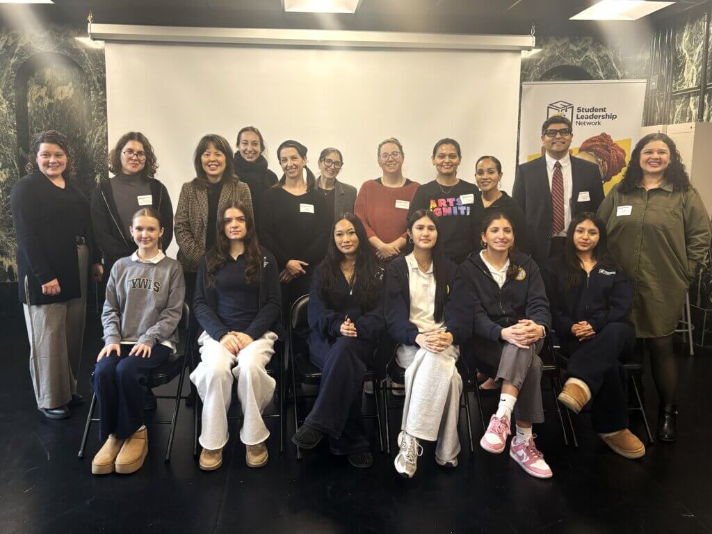 A group of students and adults pose indoors in front of a projector screen and a Student Leadership Network banner. Some are seated, while others stand behind them.