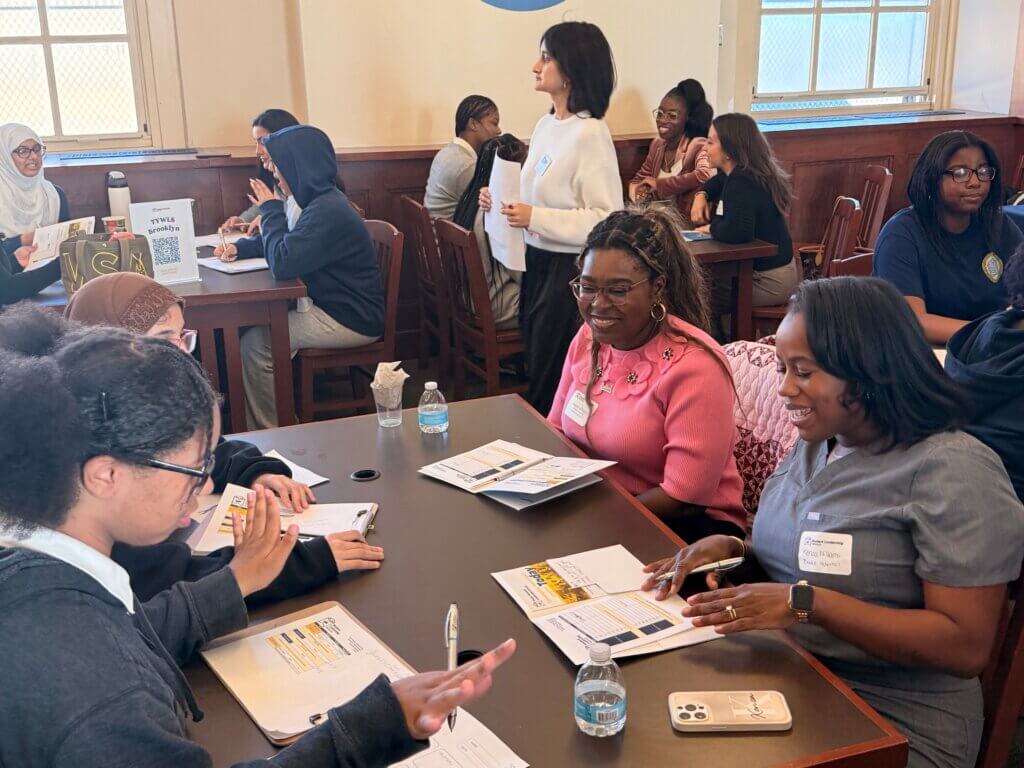 A group of students and adults sit at tables engaged in conversation, with papers, water bottles, and notebooks on the tables in a classroom setting.