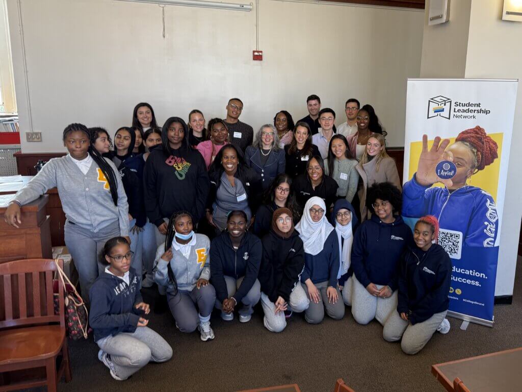 A group of students and adults pose together in a classroom near a Student Leadership Network banner. Most are wearing school uniforms.
