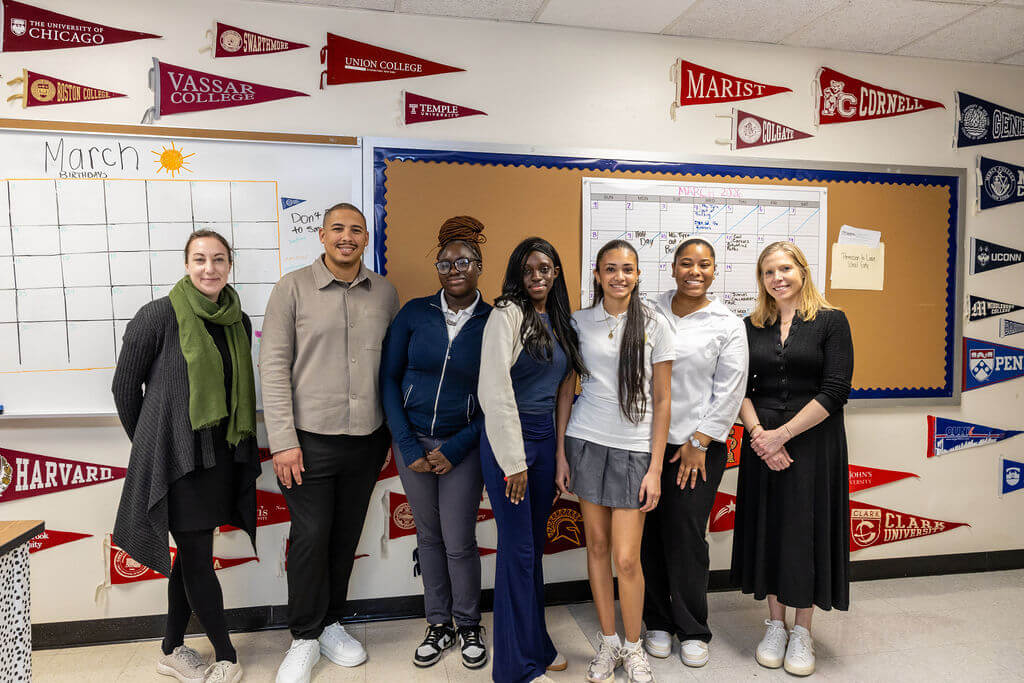 A group of seven people stands in a classroom in front of college pennants and a bulletin board, posing for a photo.