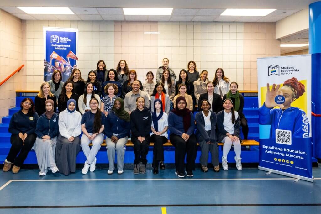 A group of adults and students pose together on bleachers in a gym, with a “Student Leadership Network” banner displayed on the right side.