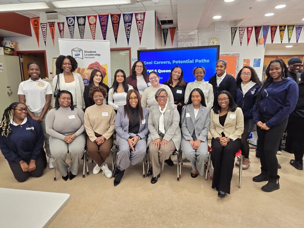 A group of women and girls pose together in a classroom, with college banners on the walls and a presentation screen in the background.