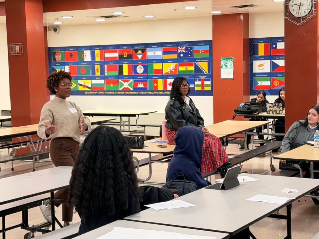 Two women address a group seated at cafeteria tables; international flags are displayed on the wall behind them. Papers and a tablet are on the tables.