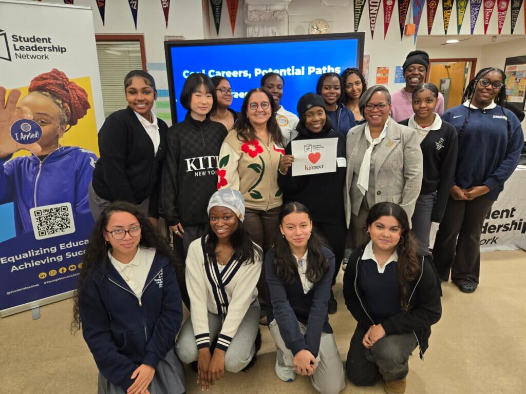 A group of students and adults pose indoors in front of a screen displaying Cool Careers, Potential Paths. Some hold a sign with a heart and the word Kindness.