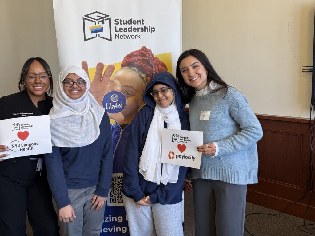 Four young women stand together smiling, two holding signs for NYU Langone Health and Paylocity, in front of a Student Leadership Network banner.