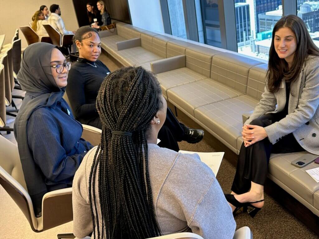 Four women sit in a modern office lounge, engaged in conversation. Papers and a smartphone are on the table nearby. Large windows overlook city buildings.