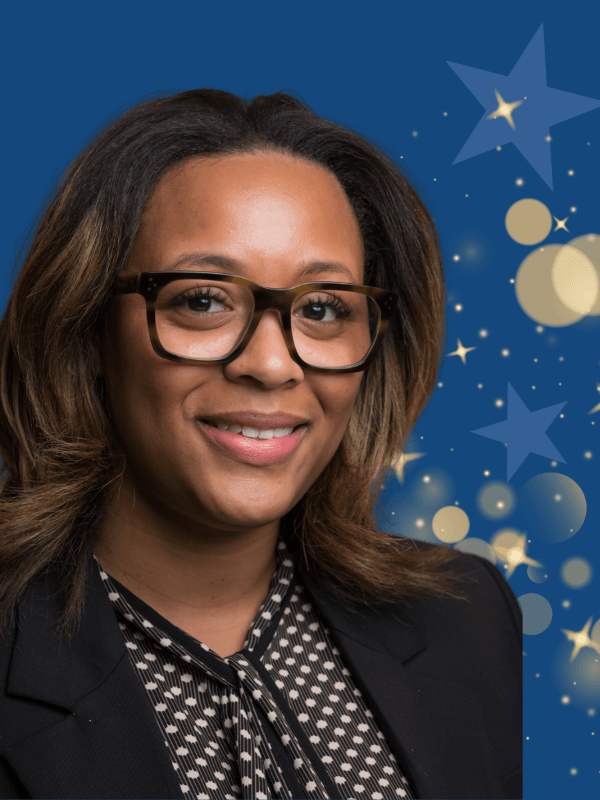 A woman with glasses and shoulder-length hair wearing a patterned blouse and blazer, smiling in front of a blue background with stars and circles.
