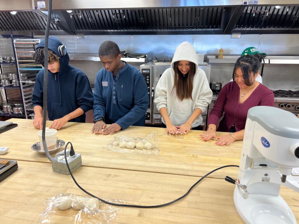 Four people stand at a kitchen counter, shaping dough balls by hand. Various kitchen equipment and utensils are visible in the background.