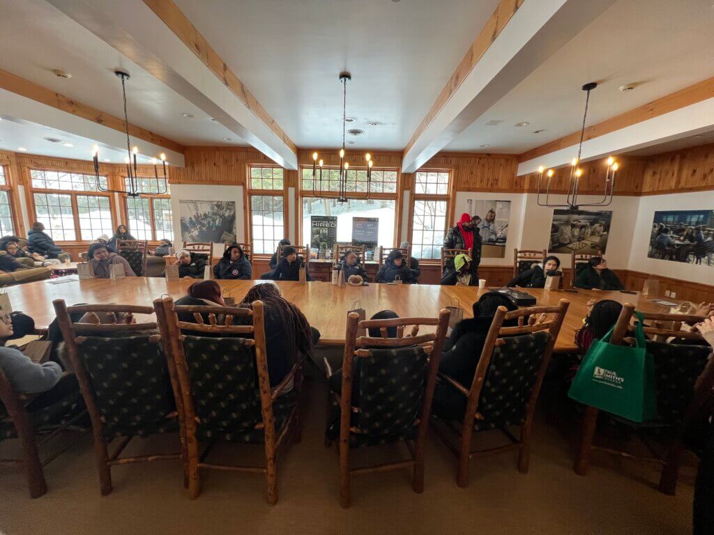 A group of people sit around a large wooden conference table in a room with wood-paneled walls and large windows, listening to someone speak.
