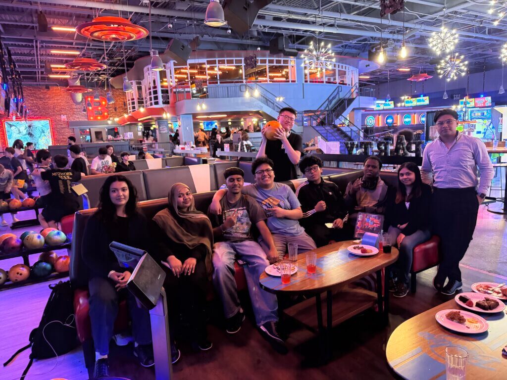 A group of nine people poses for a photo in a bowling alley lounge area, with bowling balls, food, and arcade games visible in the background.