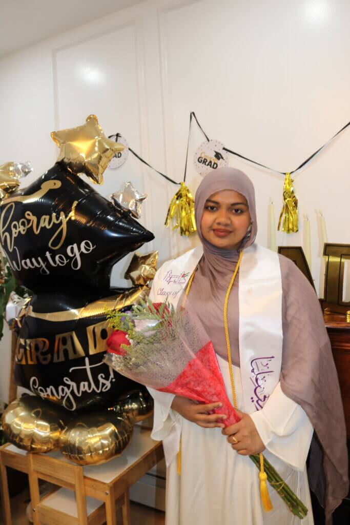 Mayisha is wearing a graduation gown and hijab holds a bouquet of flowers, standing in front of graduation-themed balloons. She will become an educator and go on to graduate school.