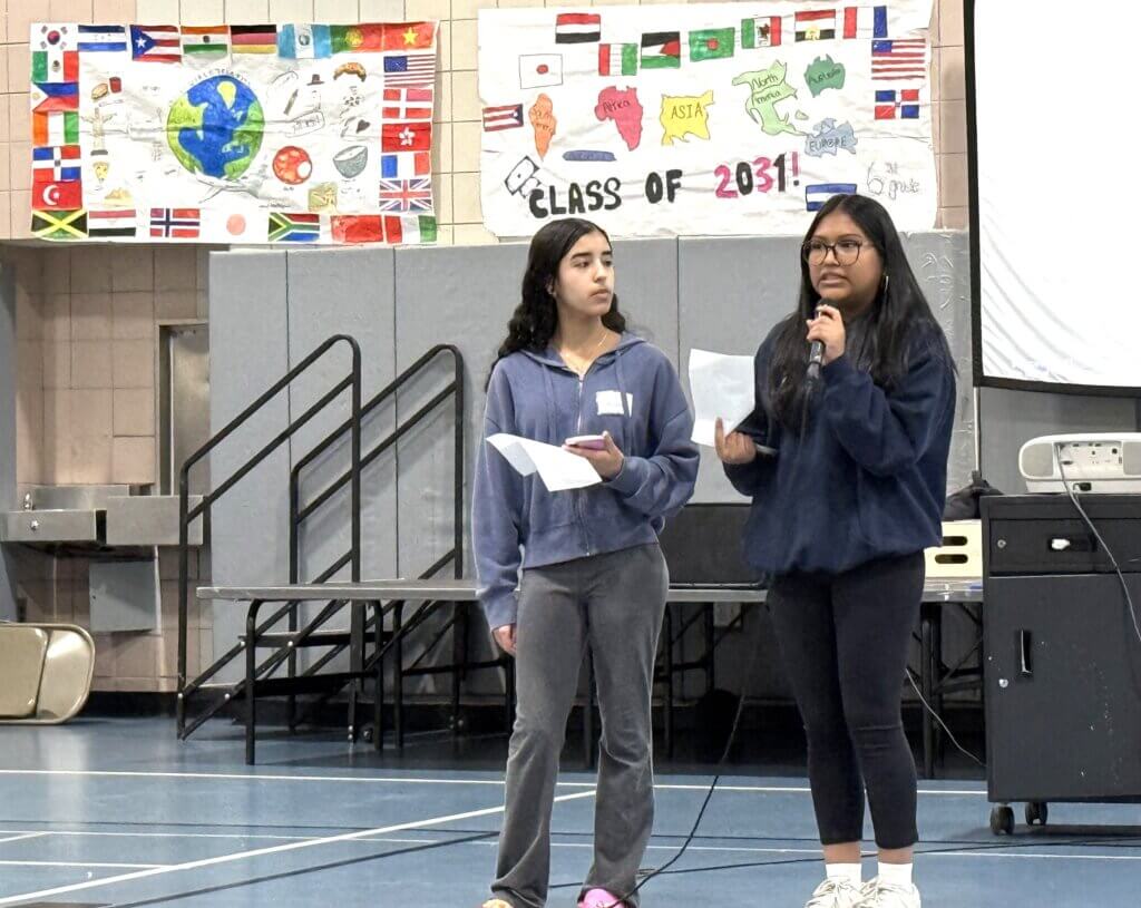 Two girls stand in a gymnasium, one holding a microphone and speaking, to give advice on scholarship applications, summer internships, and other college processes