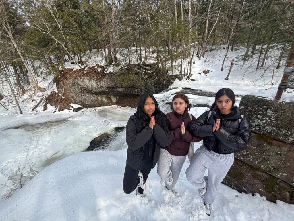 Three young women in winter clothing stand on snow, making a yoga gesture with their hands