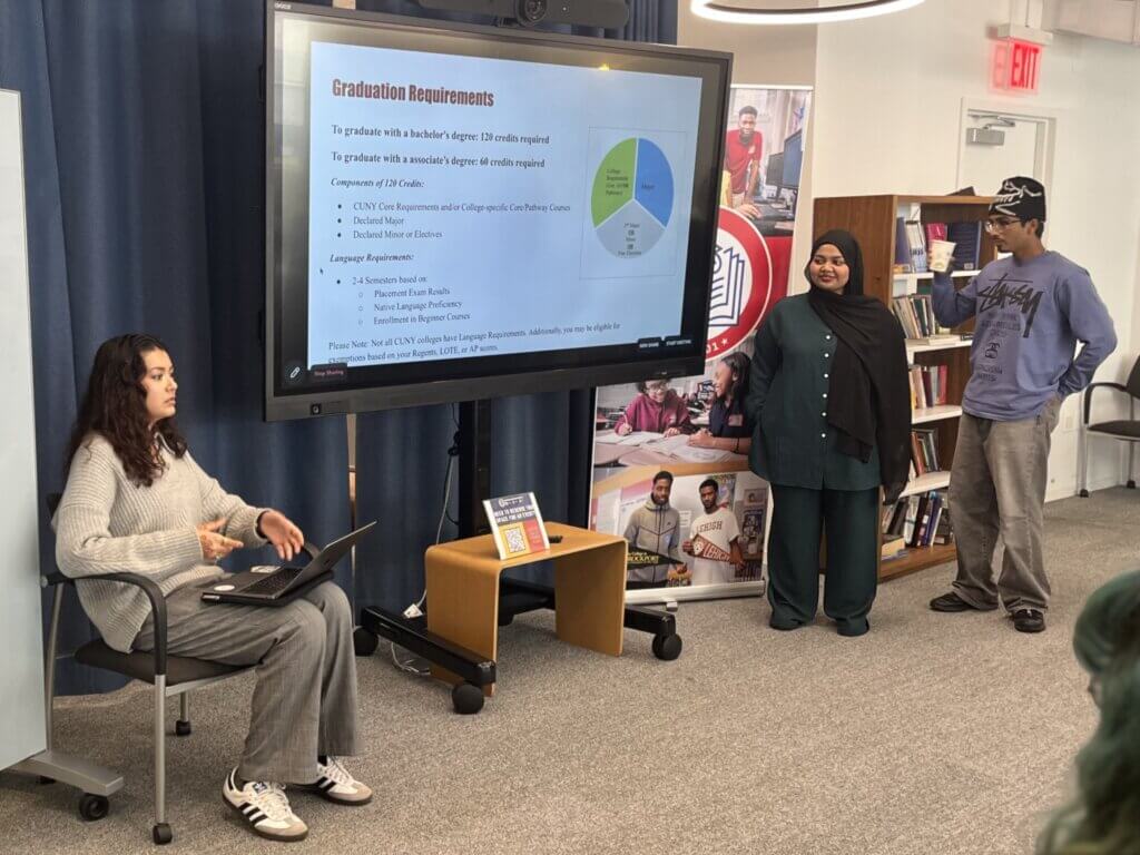 Three people present graduation requirements using a slideshow in a classroom—one seated at a laptop and two standing near a display board—illustrating the journey from student to teacher or the start of a teaching career for any young educator.