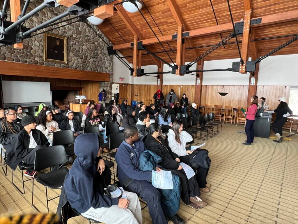 A group of student on a college trip are in a spacious room with wooden beams, stone wall, and rows of chairs at Paul Smith's College