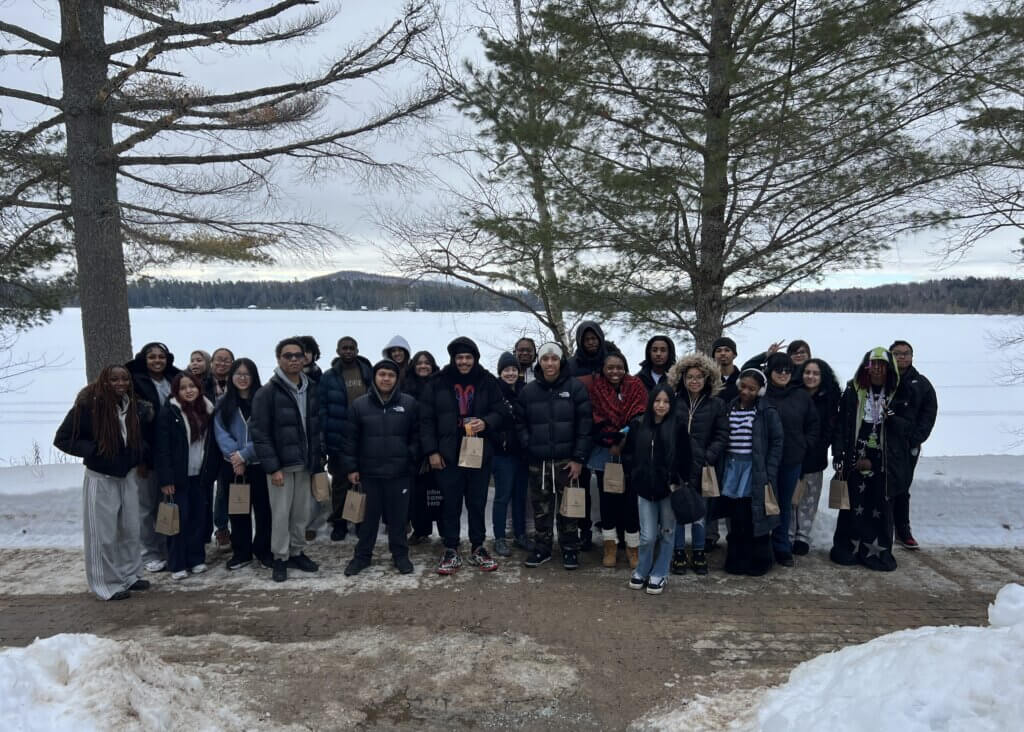 A group of students on a college trip pose for a photo outside on a snowy lakeshore, standing in front of trees with a frozen lake and forested hills in the background of Paul Smith's College campus