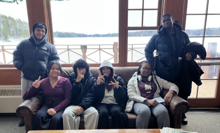 Six people sit and stand around a couch inside a cabin with large windows showing a snowy landscape and lake outside for the Paul Smith College trip