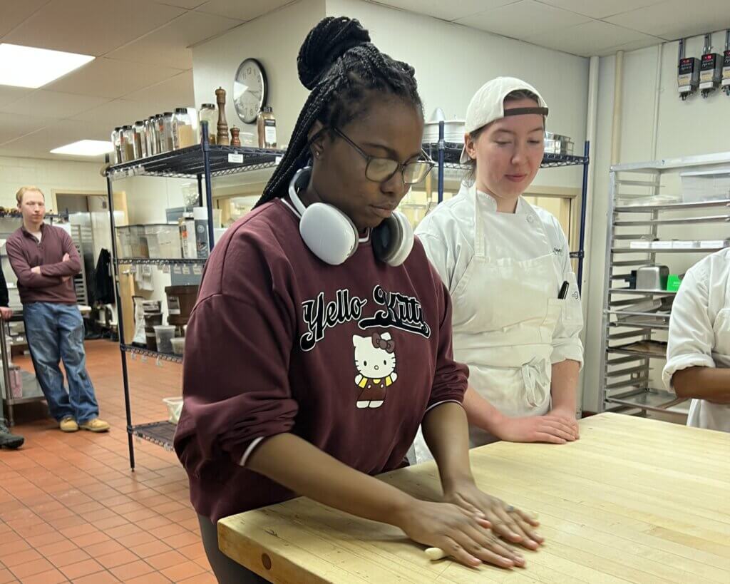A person wearing a Hello Kitty sweatshirt and headphones kneads dough on a wooden table, while a person in a white chef uniform observes in a kitchen setting.