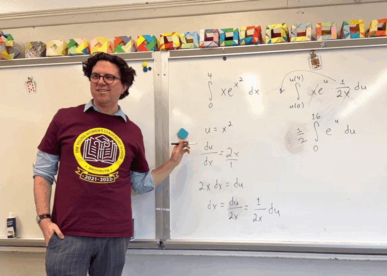 A teacher in a maroon t-shirt stands by a whiteboard filled with AP Calculus integrals and equations, holding a blue marker. Colorful paper cubes are displayed above the board at TYWLS Brooklyn.