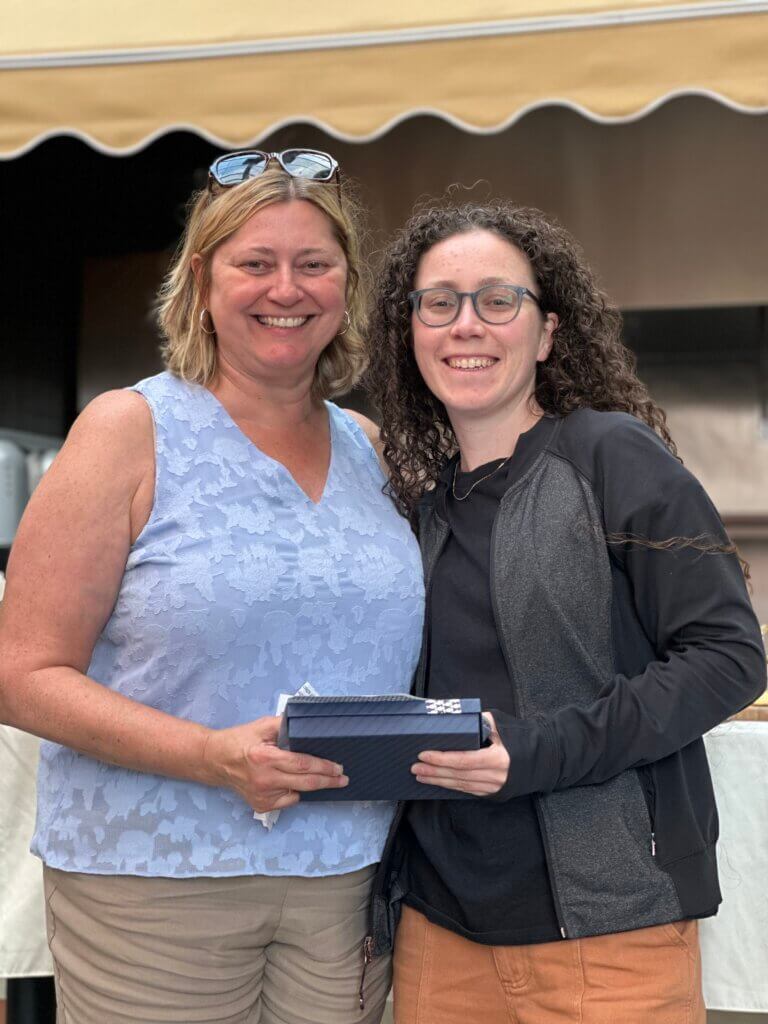 Two people stand side by side outdoors, both smiling at the network's April Blount Awards. One is holding a rectangular blue box.