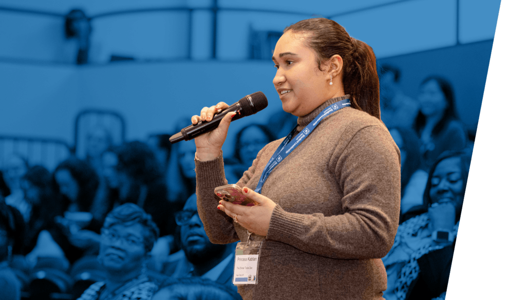 A woman speaks into a microphone while holding a phone at a Professional Learning Community event, wearing a name badge and lanyard; audience members are seated in the background.