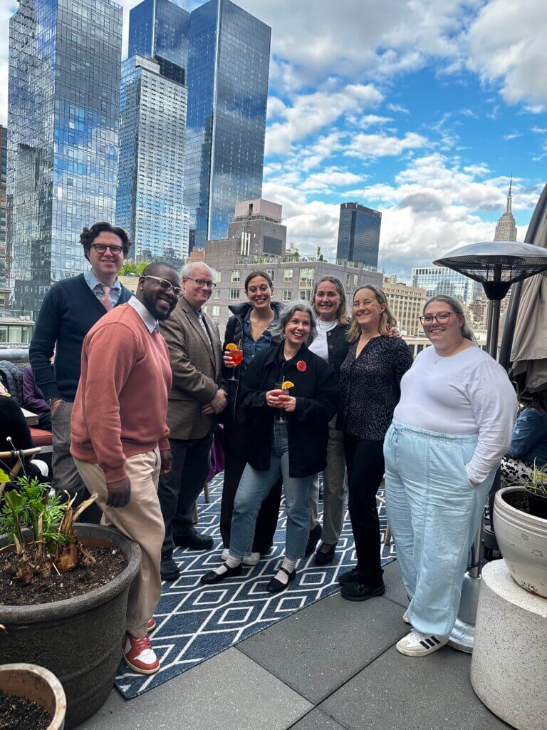 Eight people pose and smile on a city rooftop with tall buildings and a partly cloudy sky in the background for the network's April Blount Awards