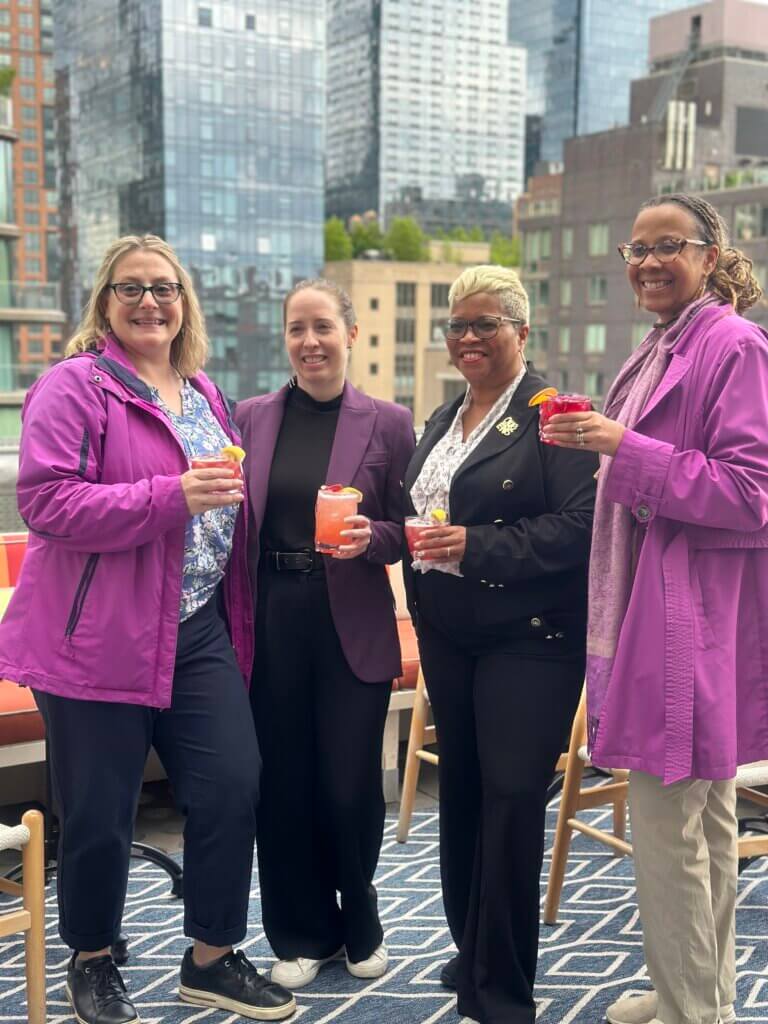 Four women stand together on an outdoor terrace, smiling and holding drinks, with tall city buildings in the background.