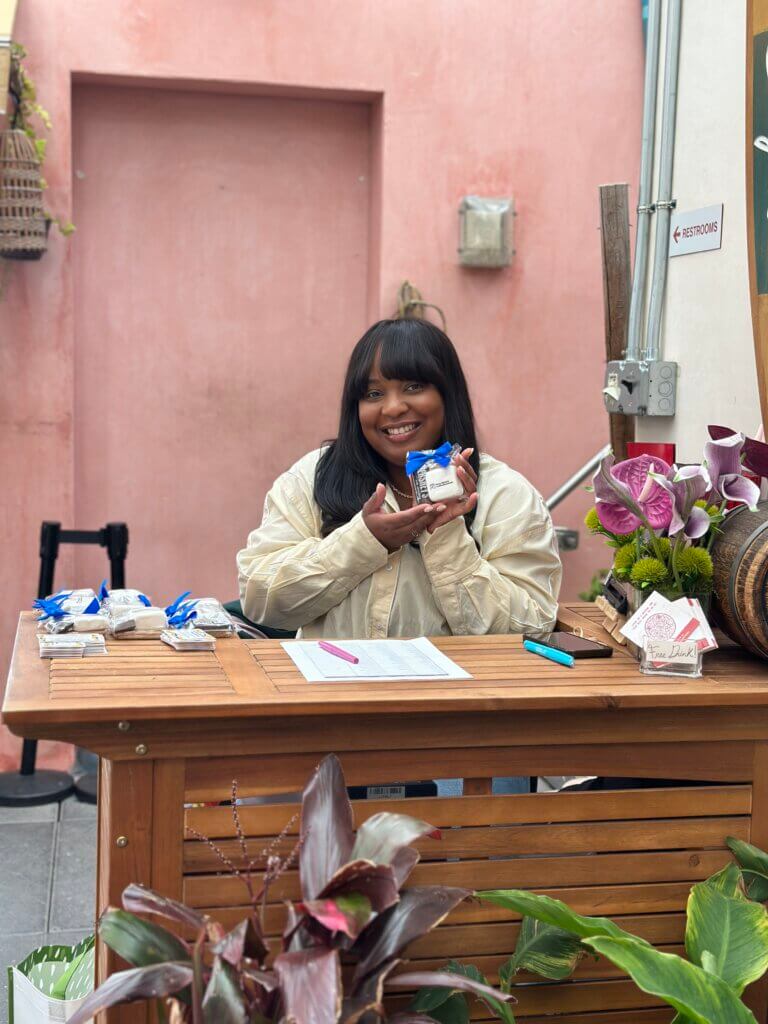 A woman sits at a wooden table, smiling and holding a packaged item behind a table with sign-in sheets