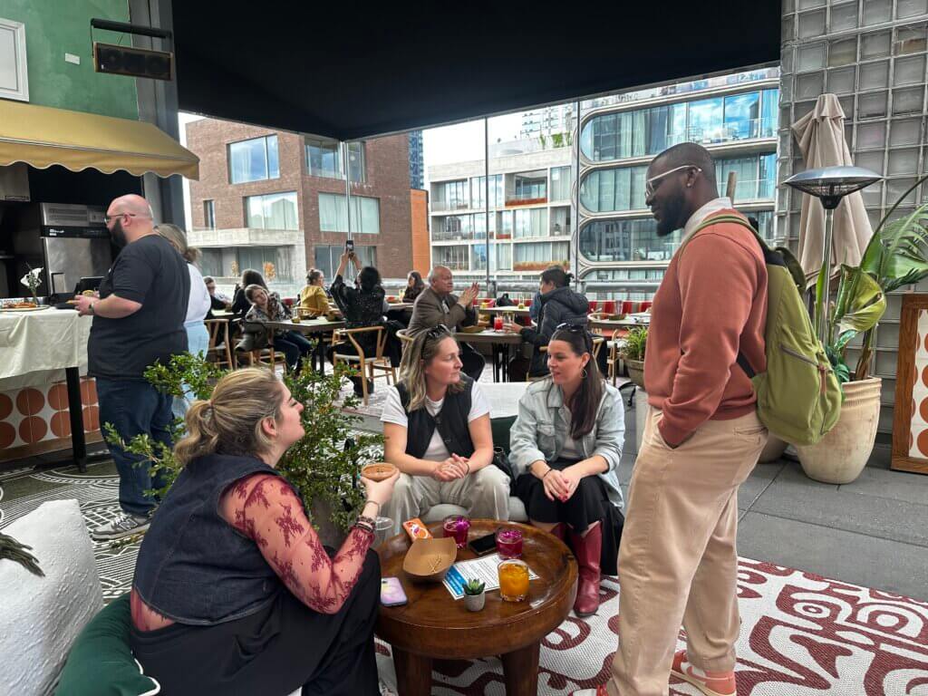 A group of people sit and stand around a low table on an outdoor patio, having conversations; other people are seated and eating in the background.