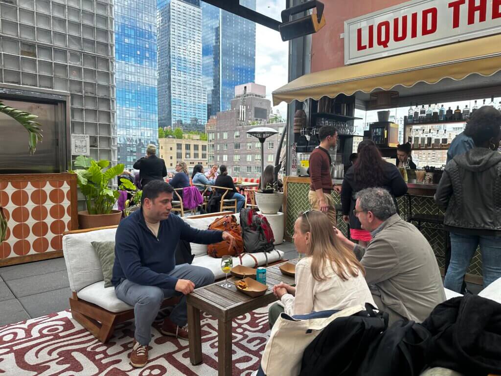 Three people sit and talk at an outdoor rooftop lounge with city buildings in the background