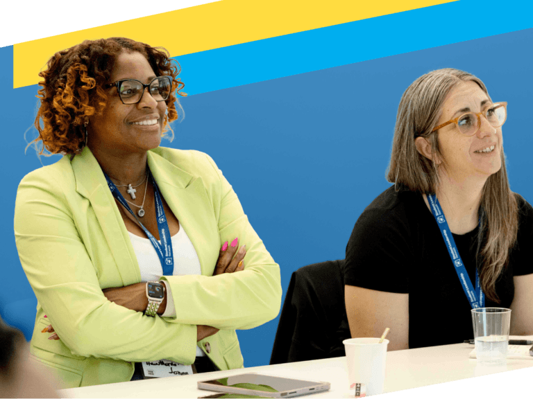 Two women sitting at a table with conference lanyards, one smiling with arms crossed, the other looking attentive; cups and electronic devices on the table—an engaging moment in a Professional Learning Community.