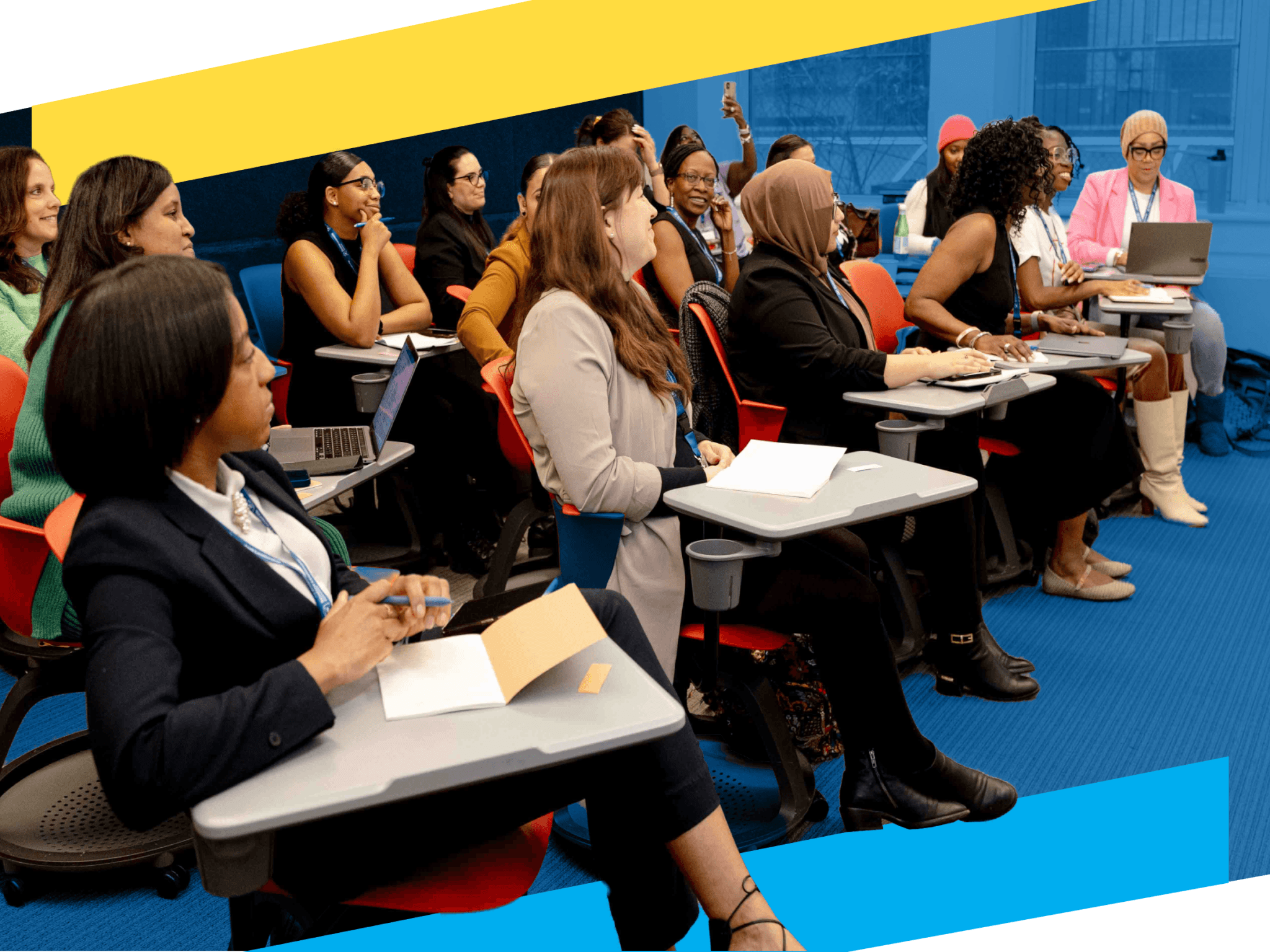 A diverse group of people sit in a classroom setting, some taking notes and others using laptops, participating in a Professional Learning Community seminar or workshop.