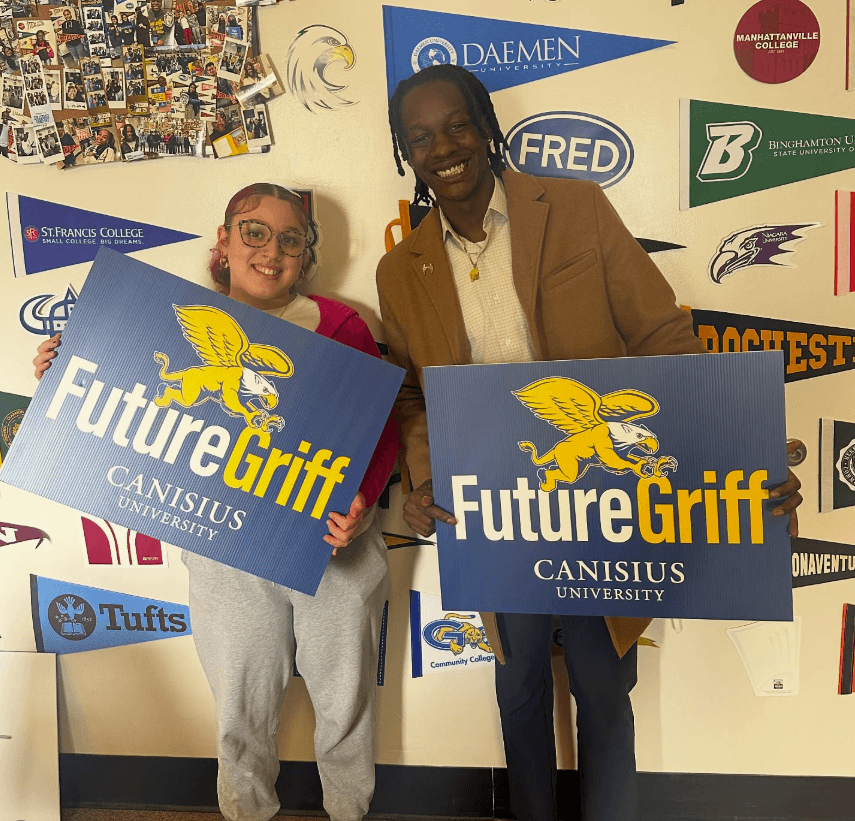 Two people stand indoors in front of a college pennant wall, each holding a Future Griff Canisius University sign with a griffin logo.