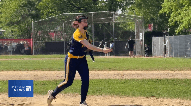 A softball pitcher in a navy and yellow uniform is mid-throw on a field, with spectators and a chain-link backstop in the background. A “Spectrum News 1” logo appears in the corner.