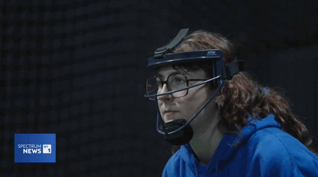 A person wearing glasses, a blue hoodie, and a protective face guard looks ahead with a focused expression on a softball field