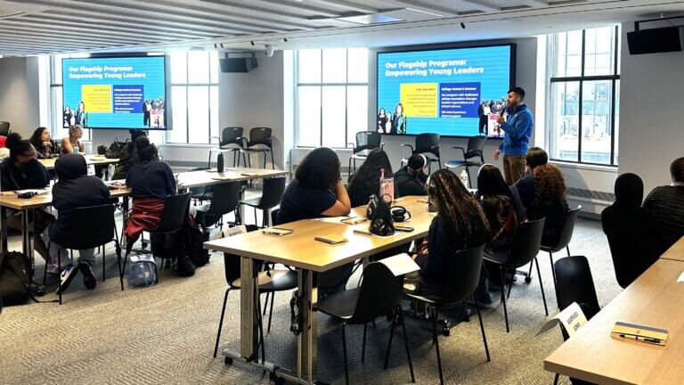 A speaker presents a slide about youth leadership programs to a group of attendees seated at tables in a conference room at Sony.