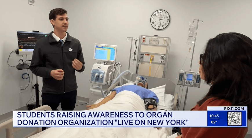 A young man speaks to a woman in a hospital room while a simulated patient lies in a hospital bed with medical equipment in the background as part of a broadcast about organ donation awareness