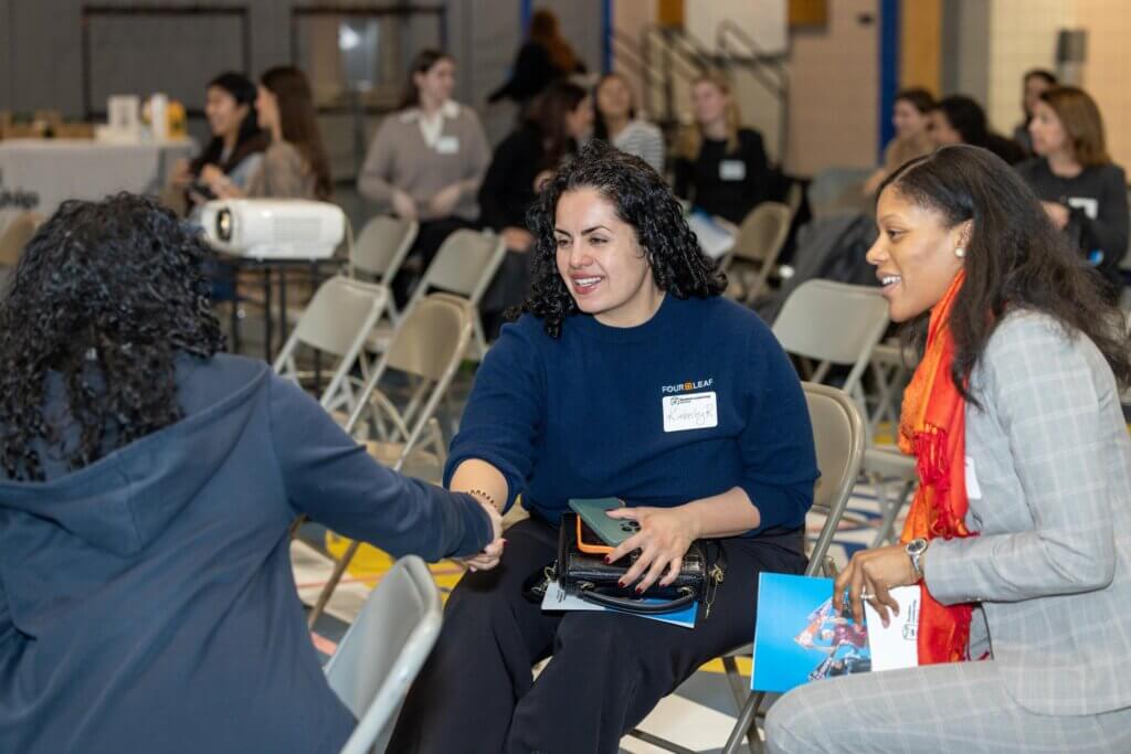 Two women and a student are seated in folding chairs at an indoor event greet each other, with one reaching out for a handshake