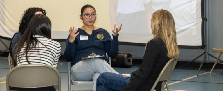 Two adults and two students sit in a circle having a discussion about early career exploration, in front of a large projected screen that reads “Let’s Practice!”