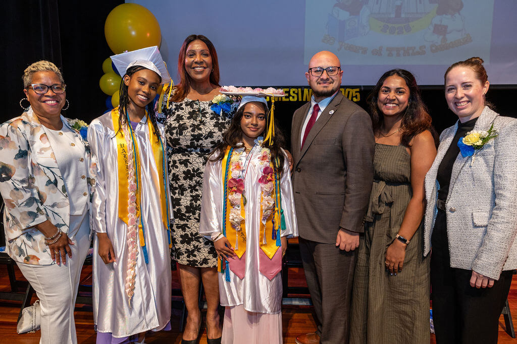 Two students in graduation caps and gowns stand on stage with their school principal, NYS Attorney General Letitia James, and other educators