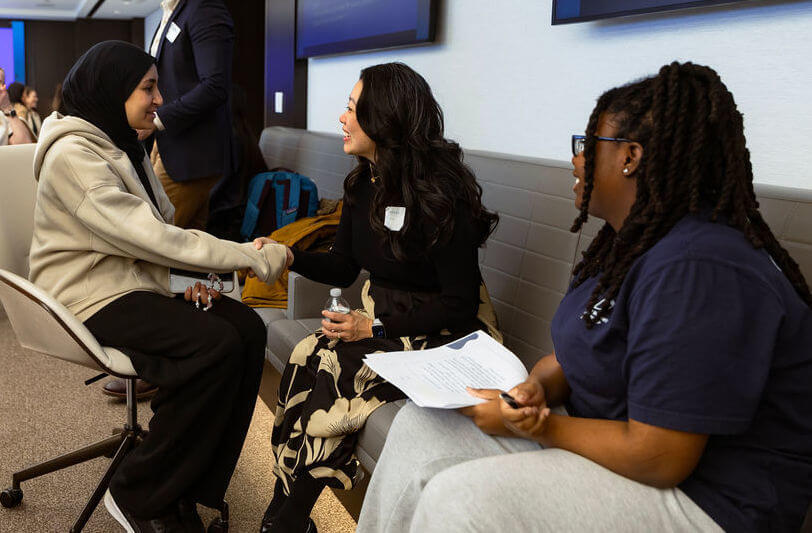 Three women seated indoors; two are shaking hands while another observes and holds papers, suggesting a discussion in a professional setting—possibly a meeting related to the Apollo Opportunity Foundation.