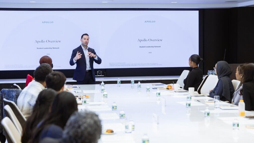 A man in a suit presents to a group seated around a conference table with “Apollo Overview” and the Apollo Opportunity Foundation displayed on two large screens behind him.