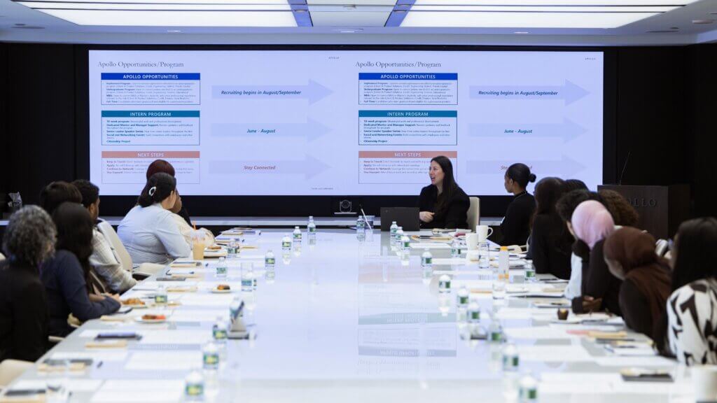 A group of people sit around a conference table listening to a presenter from the Apollo Opportunity Foundation in front of two large screens displaying program information slides.