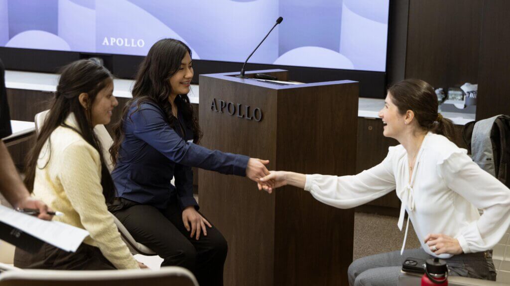 Three women in business attire sit and shake hands in a conference room with a podium labeled APOLLO, showcasing a partnership event hosted by the Apollo Opportunity Foundation, with a large screen in the background.
